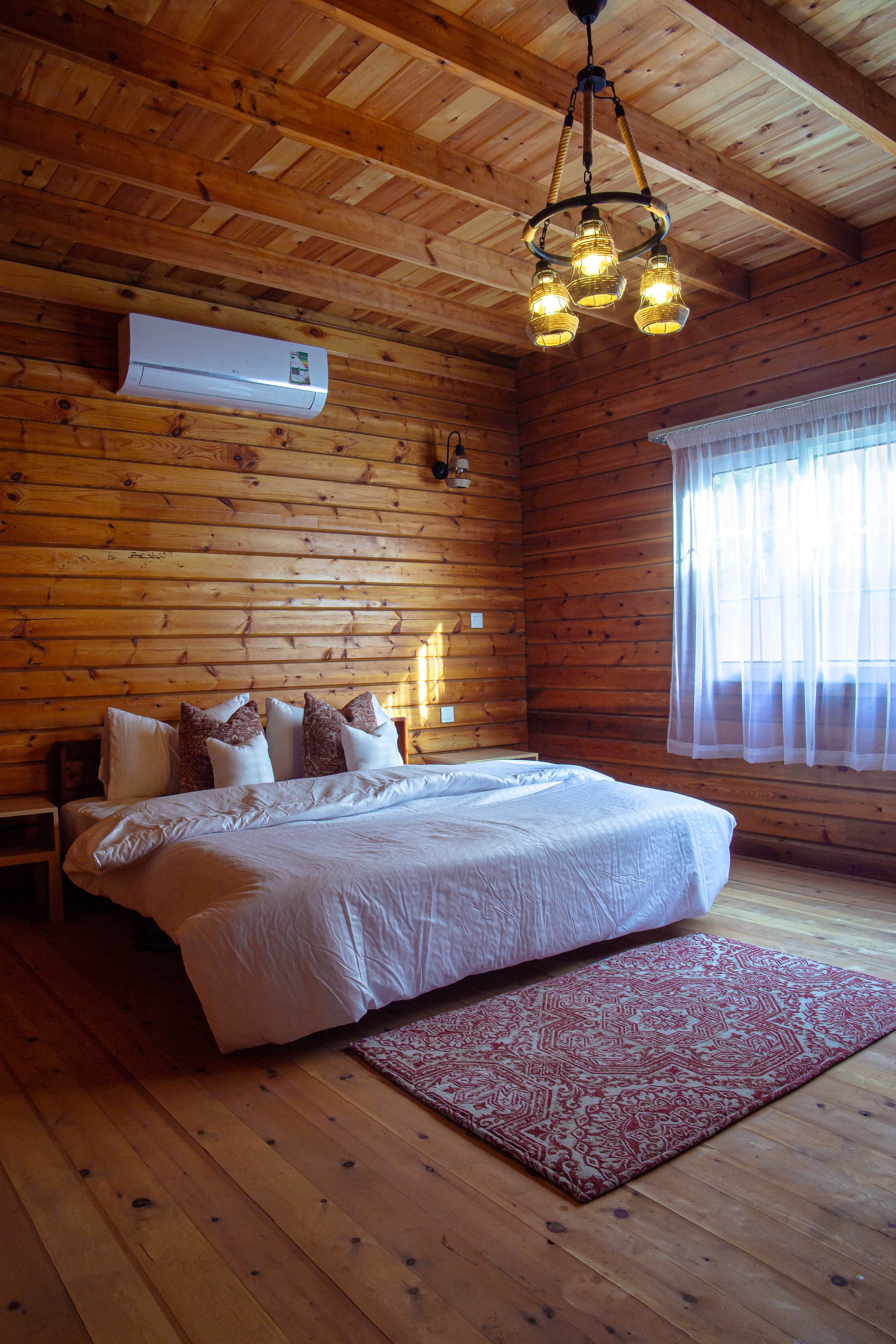 Master Bedroom log cabin with king bed white duvet single window and sheer white curtain; red patterned rug.